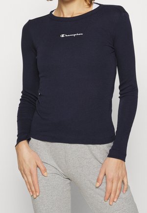 Kobieta ubrana w dopasowaną granatową koszulkę z długim rękawem z logo "Champion" na piersi oraz jasnoszare spodnie dresowe, stojąca z rękami spoczywającymi na udach.