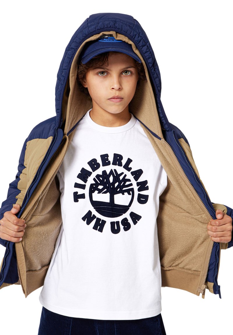 Giacca imbottita blu navy e beige aperta, che rivela una maglietta bianca con un logo scuro, raffigurante un albero e la scritta "Timberland NH USA". Tessuto simile alla lana.