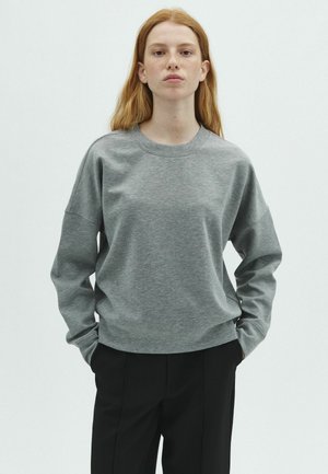 Junge Person mit langen roten Haaren, die einen grauen Pullover und schwarze Hose trägt, vor einem einfarbigen hellen Hintergrund stehend.