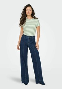 Stribet grøn og hvid t-shirt kombineret med højtaljede, brede mørkeblå jeans; modelen står imod en simpel lys baggrund.