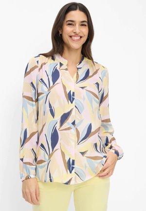 Lächelnde Frau, die eine pastellfarbene, langärmelige Bluse mit abstrakten Blattmustern und hellgelbe Hose trägt, vor einem weißen Hintergrund stehend.