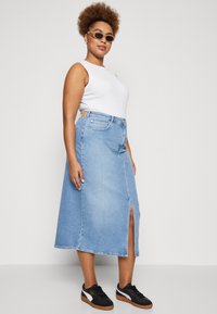 Lys blå denimskjørt med frontslitt, høy midje og lommer, kombinert med en hvit tanktop og svarte joggesko. Har en uformell design.