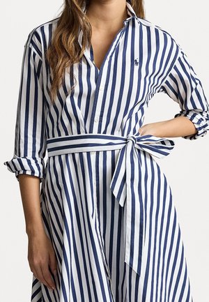 Frau trägt ein blau-weiß vertikal gestreiftes Kleid mit hochgekrempelten Ärmeln und einem gebundenen Taillengürtel.