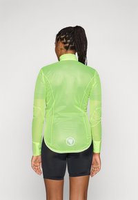 Giacca da ciclismo leggera, traslucida e a maniche lunghe di colore verde neon. Presenta un colletto alto e un logo sulla schiena, abbinata a pantaloni corti neri.