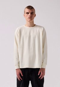 Szürkésfehér hosszú ujjú póló texturált kötött felülettel és raglán ujjal. Fekete nadrággal párosítva, a fazon laza.