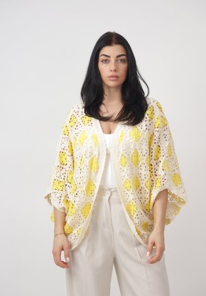 Vrouw met lang zwart haar, gekleed in een wit-gele gehaakte cardigan over een wit shirt en beige broek met hoge taille, staand tegen een effen achtergrond.