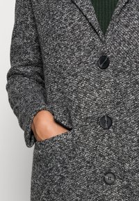 Abrigo gris texturizado con grandes botones negros, que presenta un cuello con muesca y bolsillos laterales, mostrando un patrón de tela tejido.