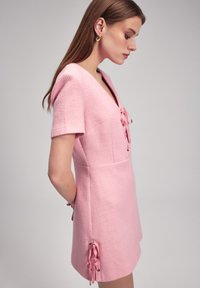 Kurzes rosa Kleid aus strukturiertem Stoff, mit V-Ausschnitt, kurzen Ärmeln und seitlichen Bändern mit Metallelementen zur Anpassung.