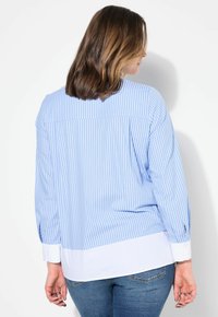 Camisa a rayas azul claro con mangas largas, puños blancos y un dobladillo inferior blanco. Presenta un yugo trasero y un ajuste holgado.