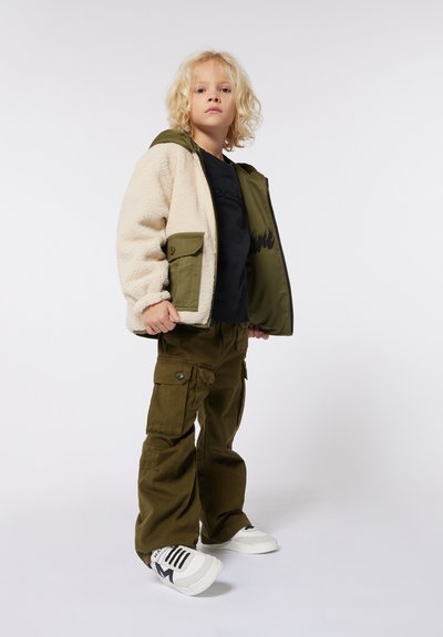 Cremefarvet fleece-foret jakke med olivengrønne accenter, sort sweatshirt, olivengrønne cargobukser og hvide sneakers med sorte striber.