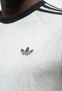 Γκρι μπλουζάκι με μαύρο γιακά και τρεις μαύρες οριζόντιες ρίγες στους ώμους. Διαθέτει κεντημένο μαύρο λογότυπο Adidas στο στήθος.