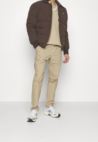 Brązowa puchowa kurtka z kołnierzem, beżowy sweter z dzianiny, jasnobeżowe spodnie typu chino oraz srebrne buty sportowe z czarnymi akcentami.