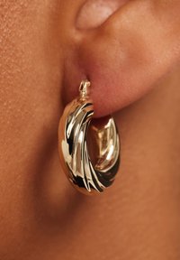 Goudkleurige oorring met een spiraalontwerp en een gepolijst oppervlak, voorzien van gestructureerde accenten en een veilige sluiting, gedragen aan de oorlel.