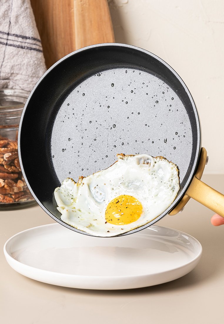 Een anti-aanbak koekenpan met een gouden handvat, gekanteld om een gebakken ei met een dooier die aan de bovenkant is, te onthullen op een wit bord.