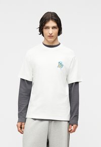 Νεαρός με μεσαίου μήκους σκούρα μαλλιά που φορά στρωτά γκρι και λευκά μακρυμάνικα και κοντομάνικα πουκάμισα, με ένα μικρό μπλε καρτούν χελώνα στο στήθος.