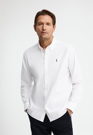 Man draagt een wit overhemd met knopen en een klein zwart logo op de borst, staand met handen in de zakken tegen een effen achtergrond.