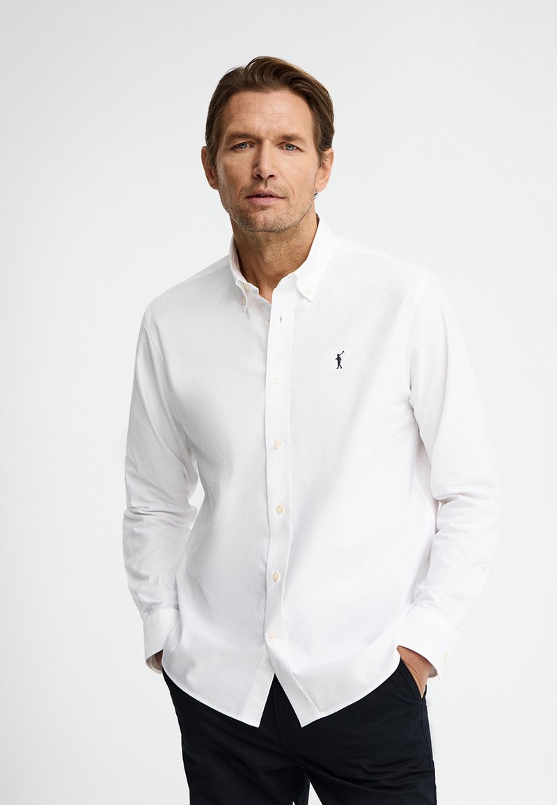 Man draagt een wit overhemd met knopen en een klein zwart logo op de borst, staand met handen in de zakken tegen een effen achtergrond.