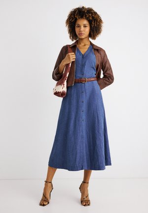 Vrouw staand, gekleed in een midi-jurk met blauwe knopen, met een bruine gevlochten riem, een bruine leren jas, patroonhakken en een bruine-witte schoudertas.