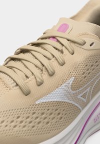 Μπεζ αθλητικό παπούτσι με λευκό λογότυπο Mizuno, ροζ λεπτομέρεια στη γλώσσα, επάνω μέρος από διαπνέον δίχτυ και μπεζ κορδόνια δεμένα πάνω από τη γλώσσα.