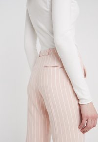 Pantalon à rayures rose clair avec une coupe ajustée, comprenant une poche arrière dissimulée et une texture de tissu lisse. Associé à un haut blanc ajusté.
