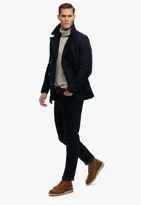 Man loopt en draagt een marineblauwe pea coat, een beige coltrui, zwarte broek, bruine laarzen en een bruine riem, tegen een effen witte achtergrond.