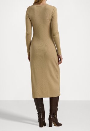 Femme debout, tournée de dos, portant une robe midi beige à manches longues et des bottes à talons hautes brun foncé.