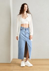 Weißes, transparentes Hemd über einem weißen Crop-Top und einem blauen Jeans-Maxi-Rock mit einem Schlitz vorne, kombiniert mit weißen Sneakers.