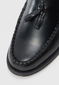 Black leather shoe with a smooth finish, featuring a stitched design and tassel detail. Rounded toe with contrasting textured trim.