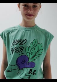 Jeune personne portant un débardeur vert avec un radis violet et le texte « Rapid Radish » imprimé à l'avant sur un fond uni.