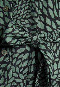 Tissu vert et noir à motif de feuilles, avec une ceinture nouée et des boutons cousus visibles sur un vêtement.