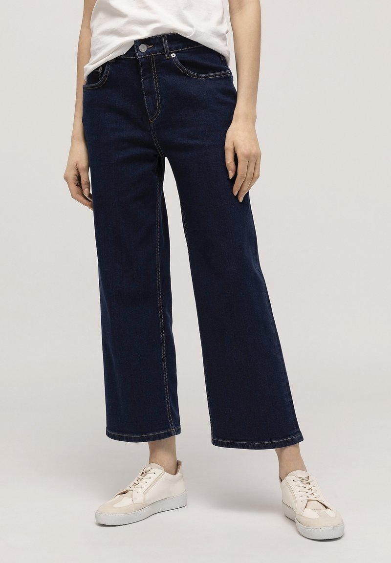 Personne portant un jean large bleu foncé, des baskets blanches et un t-shirt blanc rentré, debout devant un fond clair uni.