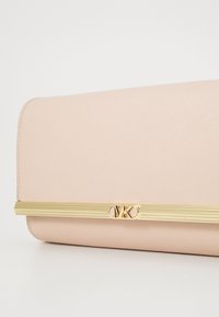 Vaaleanpunainen teksturoitu clutch-kotelo, jossa on kultasävyinen metalli-astia. Ominaisuudet sileä pinta ja vaakasuuntainen raitakoristelu, joka korostaa minimalistista muotoilua.