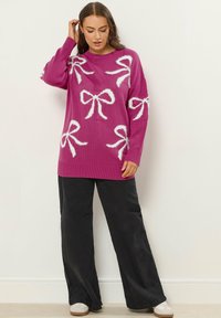 Kobieta ubrana w fuksjowy sweter w białe wzory w kokardki, czarne spodnie o szerokich nogawkach i białe trampki, stoi na tle jednolitej jasnej ściany.