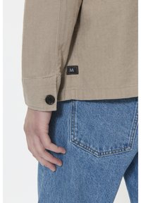 Mâneca unei jachete bej cu model herringbone, prevăzută cu un nasture negru. Etichetă "M" vizibilă pe tiv. Jeans din denim albastru deschis cu detalii la buzunarul din spate.