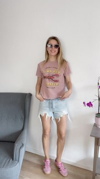 Rožinė marškinėliai su omaro grafika, šviesūs džinsiniai šortai su išradytais kraštais ir rožiniai batai. Fone matoma kėdė ir augalas.