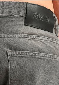 Los jeans de denim gris presentan un acabado texturizado, un patch de cuero negro en la cinturilla con "2YDNM" y costuras visibles a lo largo de los bordes.