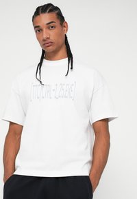 Λευκό βαμβακερό T-shirt με κοντά μανίκια, στρογγυλή λαιμόκοψη και γραφικό τύπωμα σε ανοιχτό γκρι. Χαλαρή εφαρμογή, λείος υφής, μινιμαλιστικός σχεδιασμός.