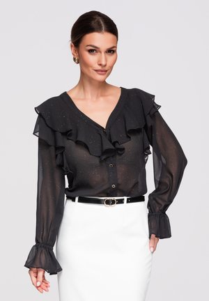 Mujer con blusa negra transparente con volantes y botones dorados, combinada con falda blanca y cinturón negro, de pie contra un fondo liso.