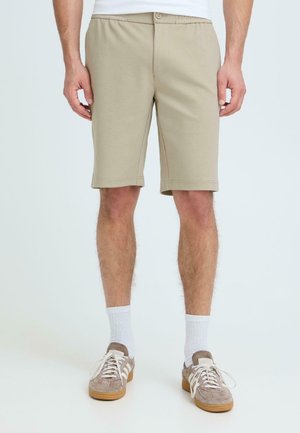 Hombre con pantalones cortos beige hasta la rodilla, calcetines blancos hasta el tobillo y zapatillas marrón claro, de pie frente a un fondo claro y liso.