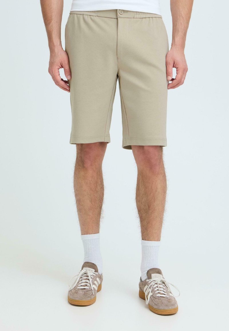 Muž v béžových krátkých šortkách po kolena, bílých ponožkách a světle hnědých teniskách stojí před jednoduchým světlým pozadím.