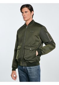 Grüne Bomberjacke mit Reißverschluss, zwei großen Fronttaschen, gerippten Bündchen und einem gestickten Logo auf der Brust. Glatte Textur.