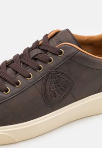 Bruna lädersneakers med bruna snören, guldfärgad öljett och en beige accent vid kragen. Har en sköldlogotyp på sidan och en texturerad yta.