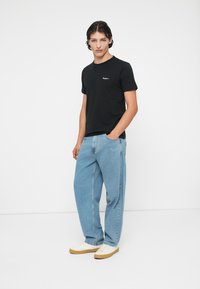 Tricou din bumbac negru, cu mâneci scurte și un mic logo alb, purtat cu blugi deschiși la culoare, cu o croială relaxată, și teniși albi cu talpă bej.