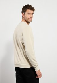 Lichtbeige sweatshirt met lange mouwen, ribbelmanchetten en -zoom, zachte textuur, voorzien van een ronde hals. Zwarte broek gedragen.