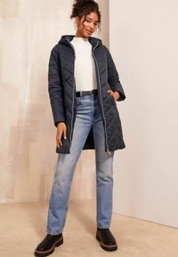 Manteau long matelassé marine avec capuche, doté d'une fermeture éclair à l'avant, de poches latérales et d'une taille cintrée, assorti à un jean bleu clair et des bottes noires.