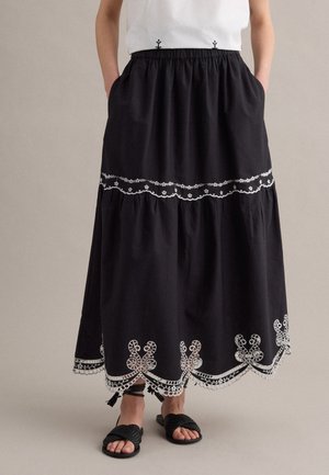 Person trägt einen schwarzen Midirock mit weißer Blumenstickerei, gewelltem Saum und Vordertaschen, kombiniert mit schwarzen gewebten Sandalen.
