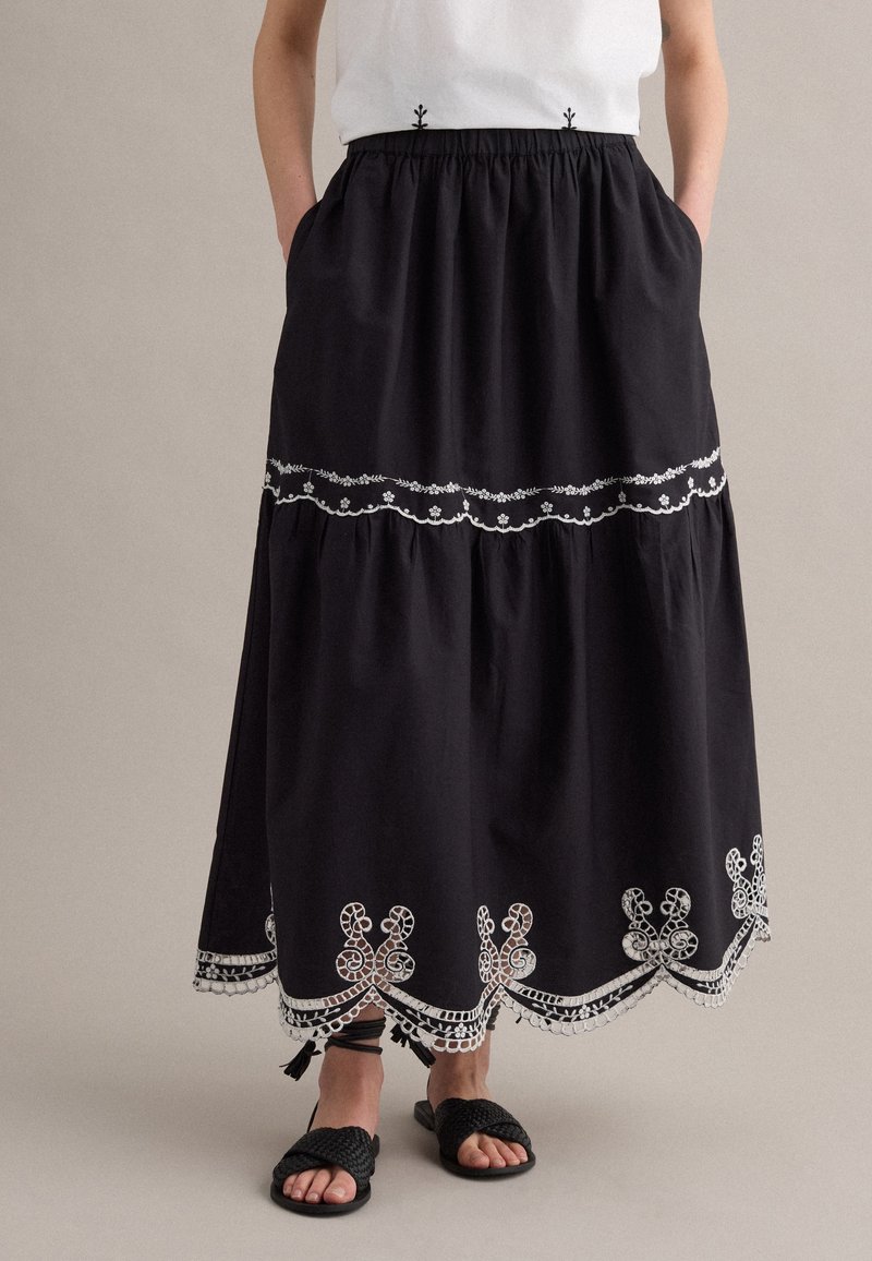 Persona que lleva una falda midi negra con bordado floral blanco, dobladillo festoneado y bolsillos delanteros, combinada con sandalias negras tejidas.