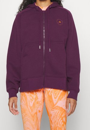Person iført en lilla hættetrøje med lynlås og forlommer samt orange leggings med mønster, stående mod en ensfarvet baggrund.