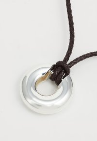 Pendente circolare in argento con superficie lucida, caratterizzato da una sezione intagliata, attaccato a una corda di pelle intrecciata di colore marrone scuro.