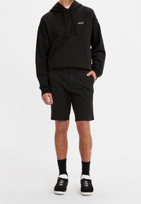 Czarna bluza z kapturem, sznurkami i kieszenią z przodu, w połączeniu z czarnymi szortami. Czarne sneakersy z białymi akcentami i czarne skarpetki. Prosty, casualowy strój.